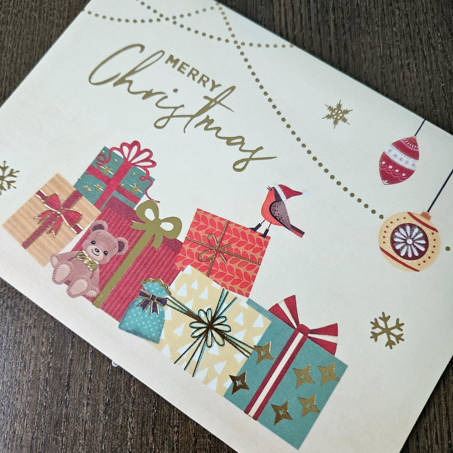 A festive Christmas greeting card featuring the words 'MERRY Christmas' in gold script, decorated with illustrated wrapped presents, ornaments, snowflakes, a small bird, and dotted line patterns, photographed on a dark wood surface.