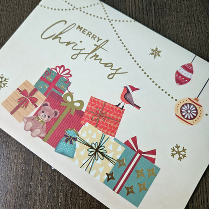 A festive Christmas greeting card featuring the words 'MERRY Christmas' in gold script, decorated with illustrated wrapped presents, ornaments, snowflakes, a small bird, and dotted line patterns, photographed on a dark wood surface.