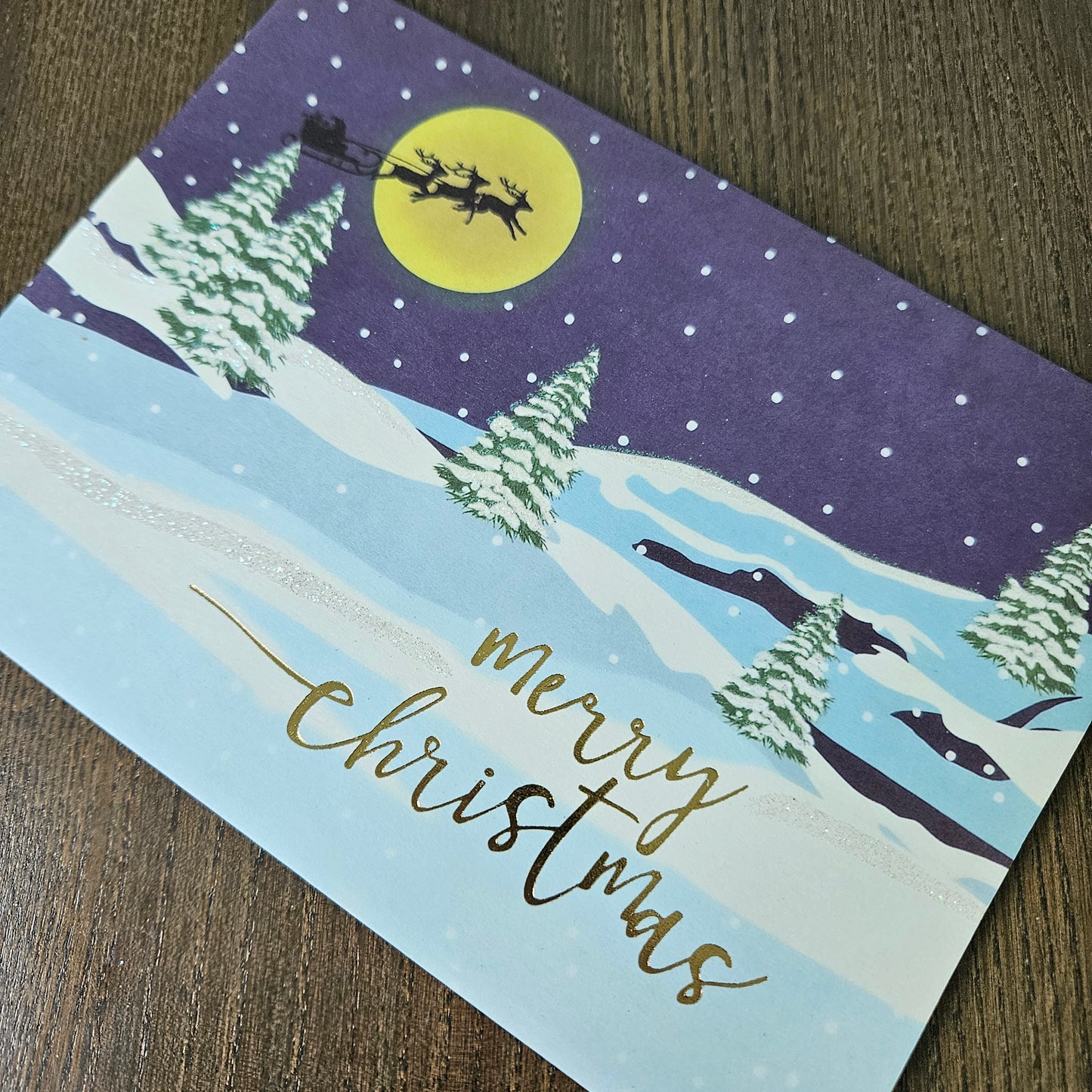A square Christmas greeting card featuring a winter night scene with Santa Claus in a sleigh under a large yellow moon, snowy mountains, pine trees, and the words 'merry christmas' in gold script, photographed on a wooden surface.
