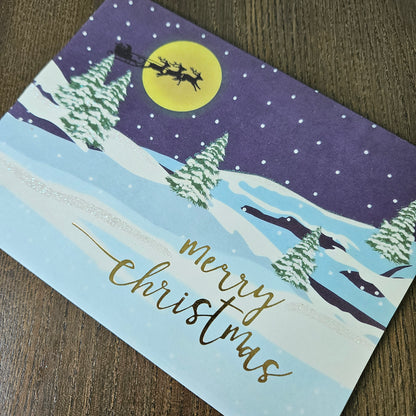 A square Christmas greeting card featuring a winter night scene with Santa Claus in a sleigh under a large yellow moon, snowy mountains, pine trees, and the words 'merry christmas' in gold script, photographed on a wooden surface.