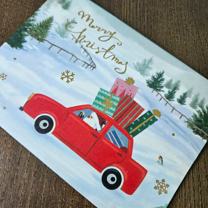 A Christmas greeting card featuring a red car carrying wrapped presents on its roof, with a Santa Claus driving, set against a snowy winter landscape. The card has gold cursive text reading 'Merry Christmas' and is photographed on a wooden surface.