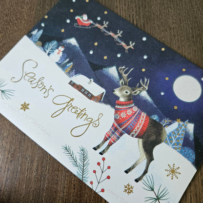 A festive Christmas greeting card lying on a wooden surface, featuring a dark blue night sky with Santa Claus in his sleigh, a deer in a red and blue sweater, a snowy house, and the words 'Season's Greetings' in gold script.