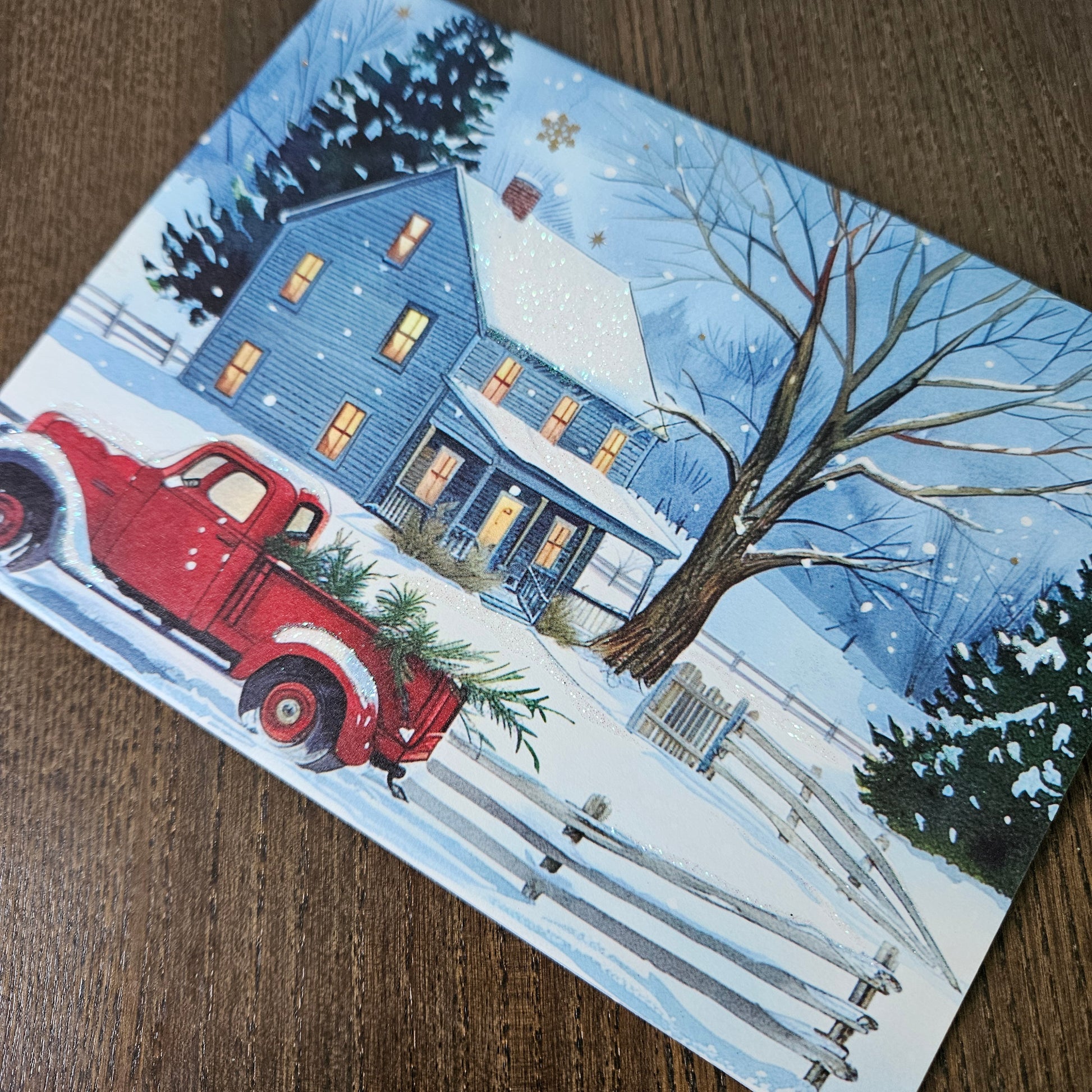 A rectangular printed card or art print featuring a winter village scene with a blue house, bare tree, snow, fence, and a red pickup truck carrying evergreen branches, photographed on a brown wood surface.