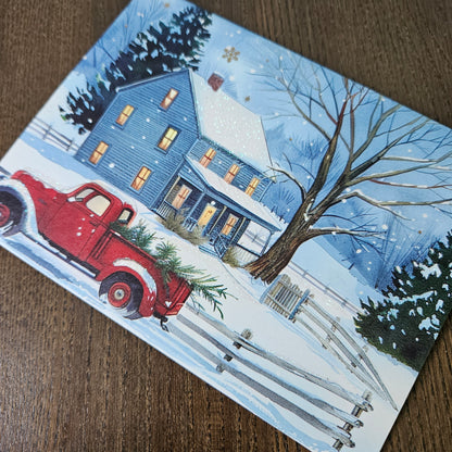 A rectangular printed card or art print featuring a winter village scene with a blue house, bare tree, snow, fence, and a red pickup truck carrying evergreen branches, photographed on a brown wood surface.