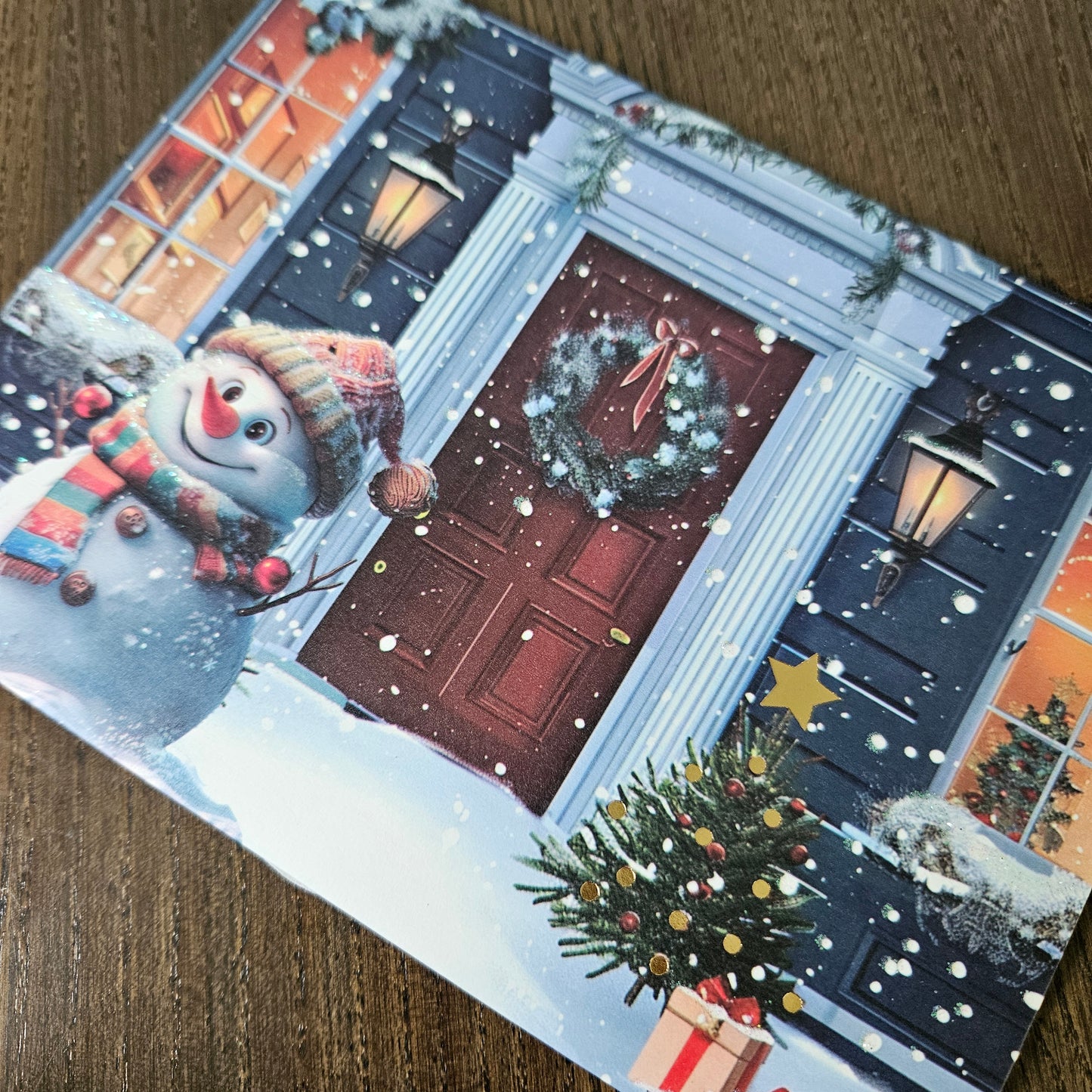 A square Christmas greeting card featuring a festive snow-covered front door with a wreath, lanterns, a snowman, and a decorated Christmas tree, photographed on a wooden surface.