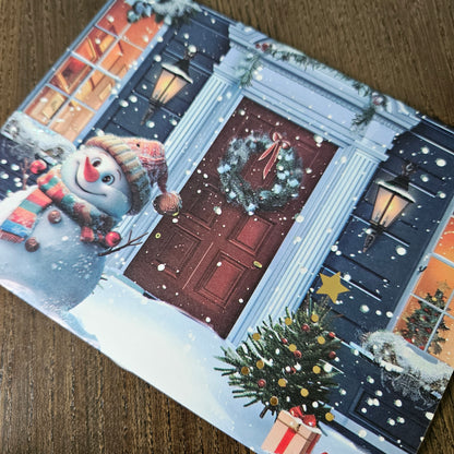 A square Christmas greeting card featuring a festive snow-covered front door with a wreath, lanterns, a snowman, and a decorated Christmas tree, photographed on a wooden surface.