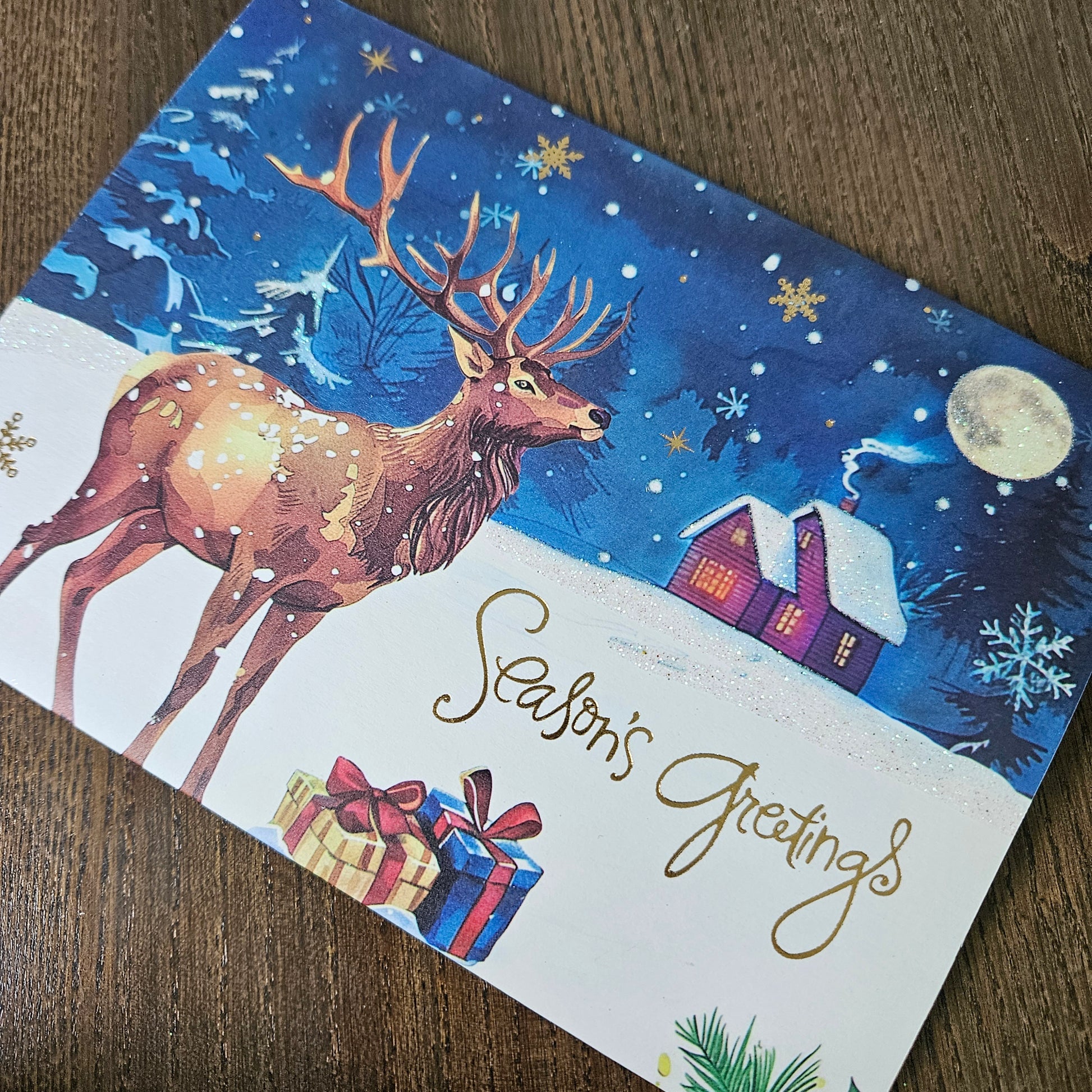 A festive Christmas greeting card featuring an illustration of a brown deer in a snowy night scene with a purple house, moon, snowflakes, and gold 'Season's Greetings' script, photographed on a wooden surface.