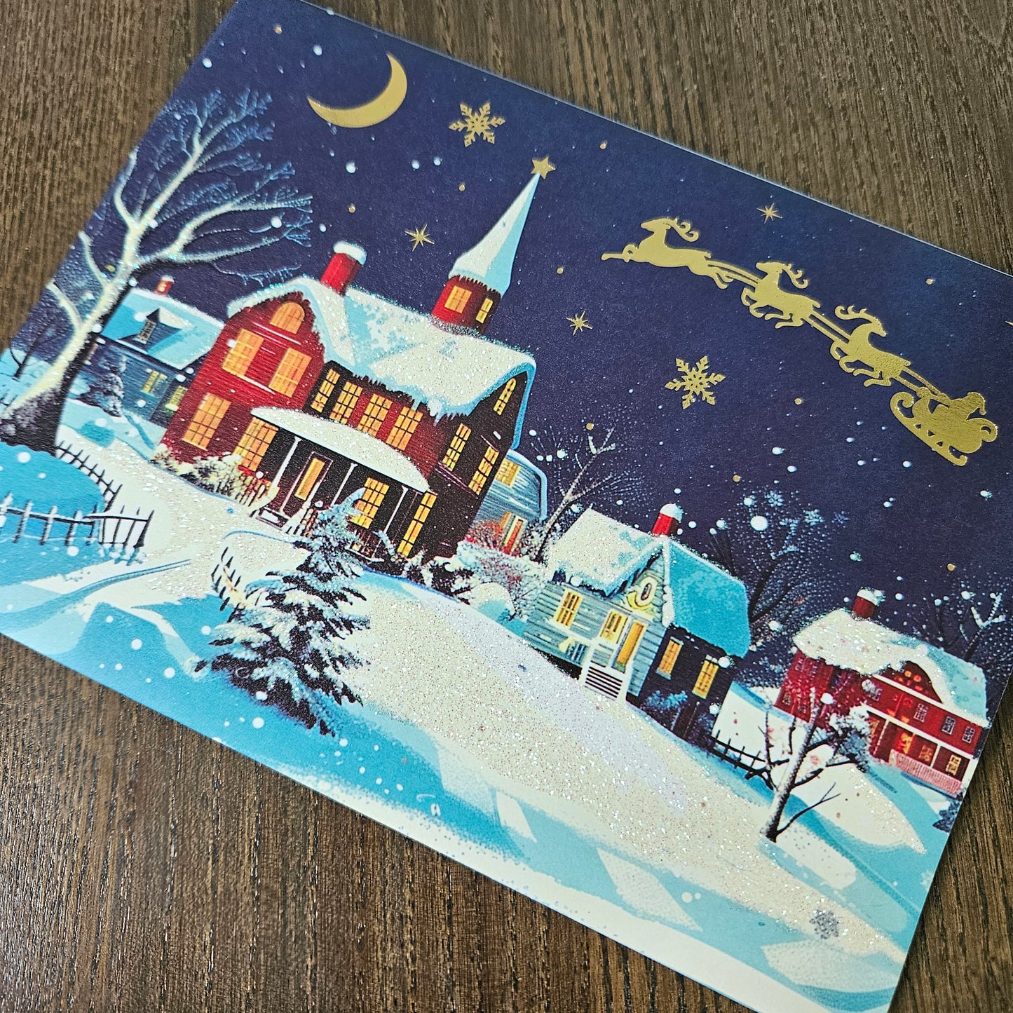 A rectangular Christmas card featuring a snowy village scene with red houses, snow-covered roofs, a crescent moon, stars, snowflakes, and Santa in his sleigh, placed on a wooden surface.