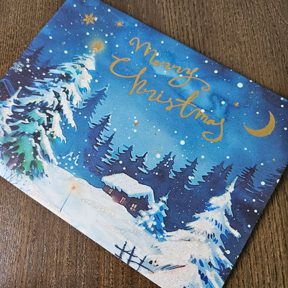 A square Christmas greeting card featuring a winter night landscape with snow-covered pine trees, a small cabin, stars, and gold foil script reading 'Merry Christmas,' photographed on a wooden surface.