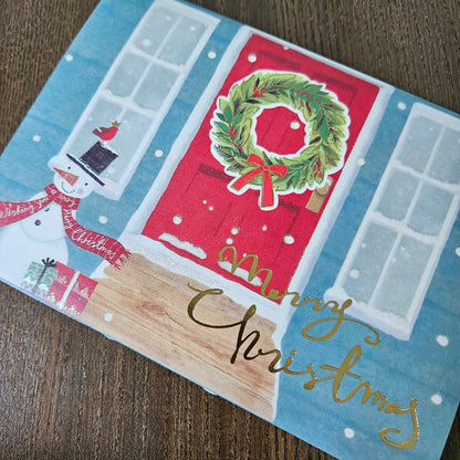 A square Christmas greeting card featuring a festive house design with a red door, green wreath, snowman, snow windows, and gold 'Merry Christmas' script, photographed on a wooden surface.