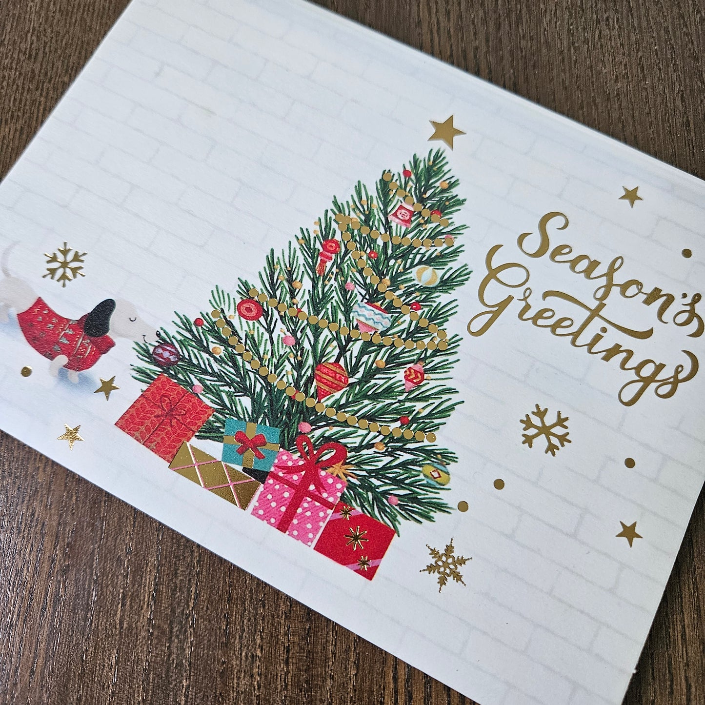 A white Christmas greeting card with a decorated evergreen tree, wrapped presents, gold stars and snowflakes, and the words 'Season's Greetings' in gold script, photographed on a wooden surface.