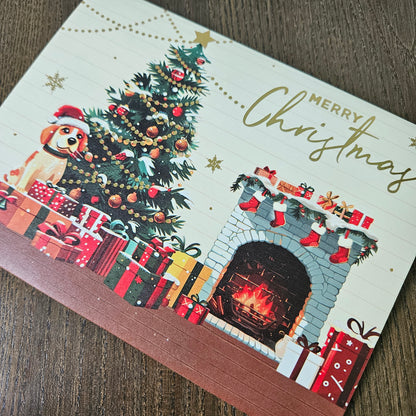 A Christmas greeting card featuring an illustrated decorated tree, gifts, a fireplace, stockings, and the words “Merry Christmas,” photographed on a wooden surface.