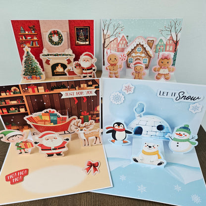 Four festive pop-up greeting cards arranged on a tabletop, each featuring holiday scenes and 3D elements: a red Christmas fireplace scene, a snowy village scene, a shelf scene with Santa and gifts, and a snowy arctic scene with penguins, a snowman, and an igloo.