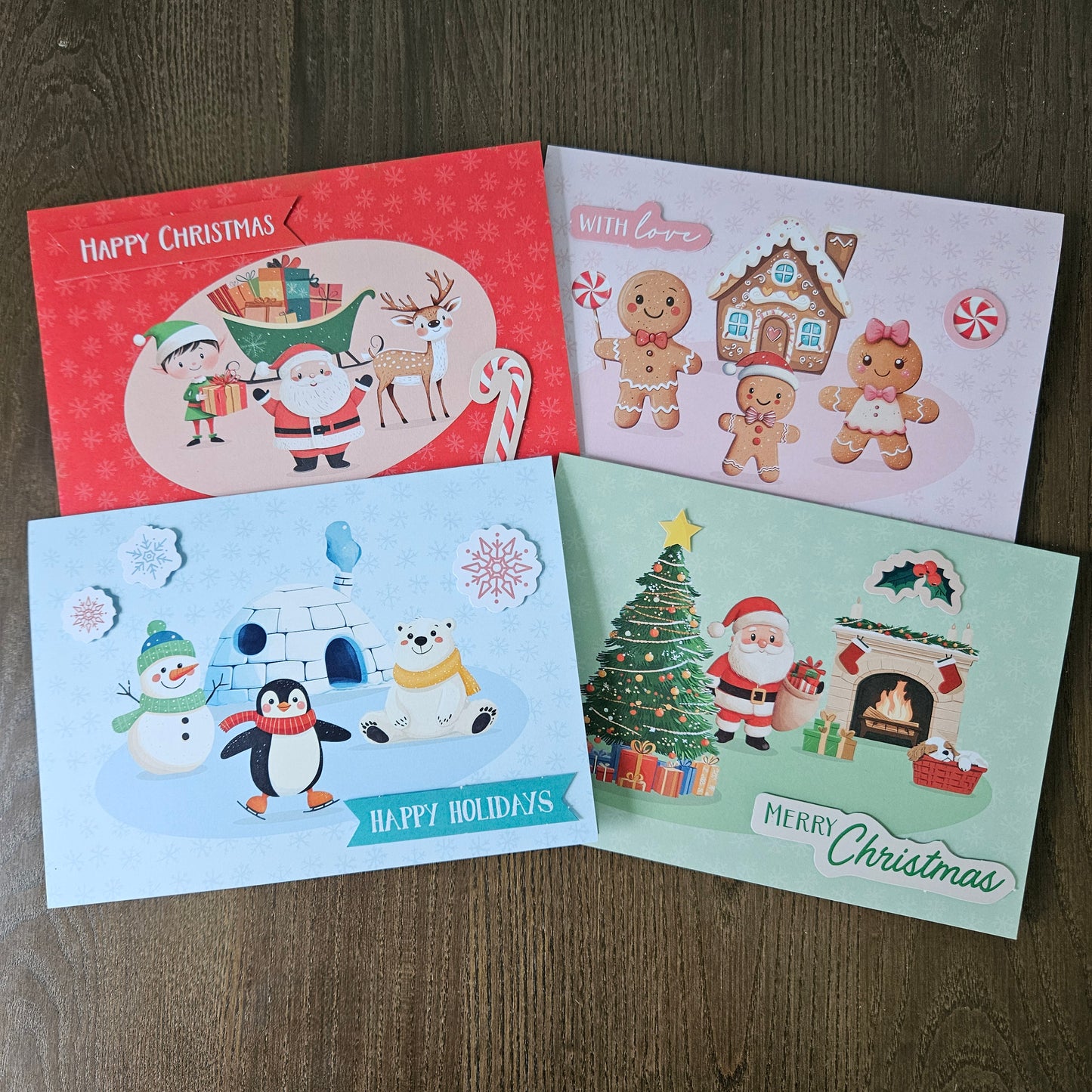 Four Christmas greeting cards arranged on a dark wood surface. The cards feature festive illustrations including Santa Claus, gingerbread people, a snowman, a polar bear, a Christmas tree, and holiday scenes, with text such as 'Happy Christmas,' 'With Love,' and 'Merry Christmas.