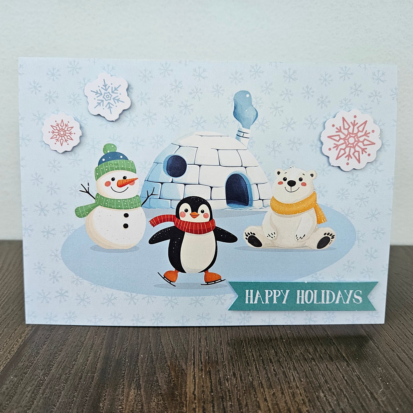 A square handmade holiday greeting card featuring a penguin, snowman, polar bear, and igloo on a snowy background, with 'HAPPY HOLIDAYS' written on a teal banner. The card is displayed on a dark wooden surface against a light gray wall.