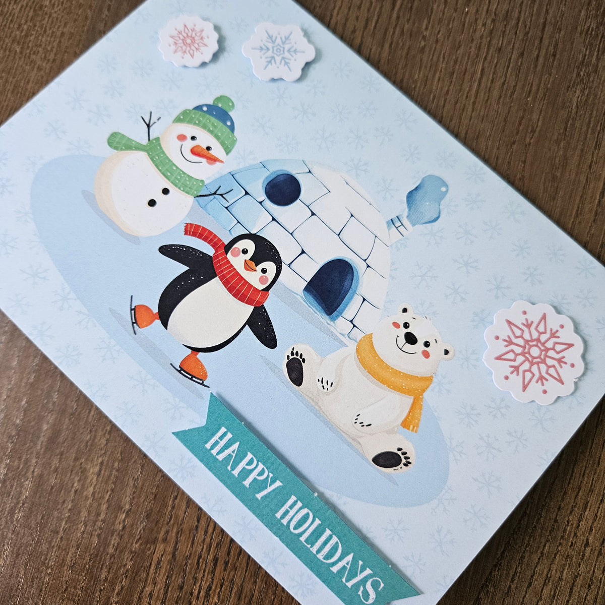 A square holiday greeting card featuring a winter scene with a penguin, snowman, polar bear, and igloo on a pale blue background, placed on a wooden surface. The card reads “HAPPY HOLIDAYS” and includes decorative snowflake stickers around it.