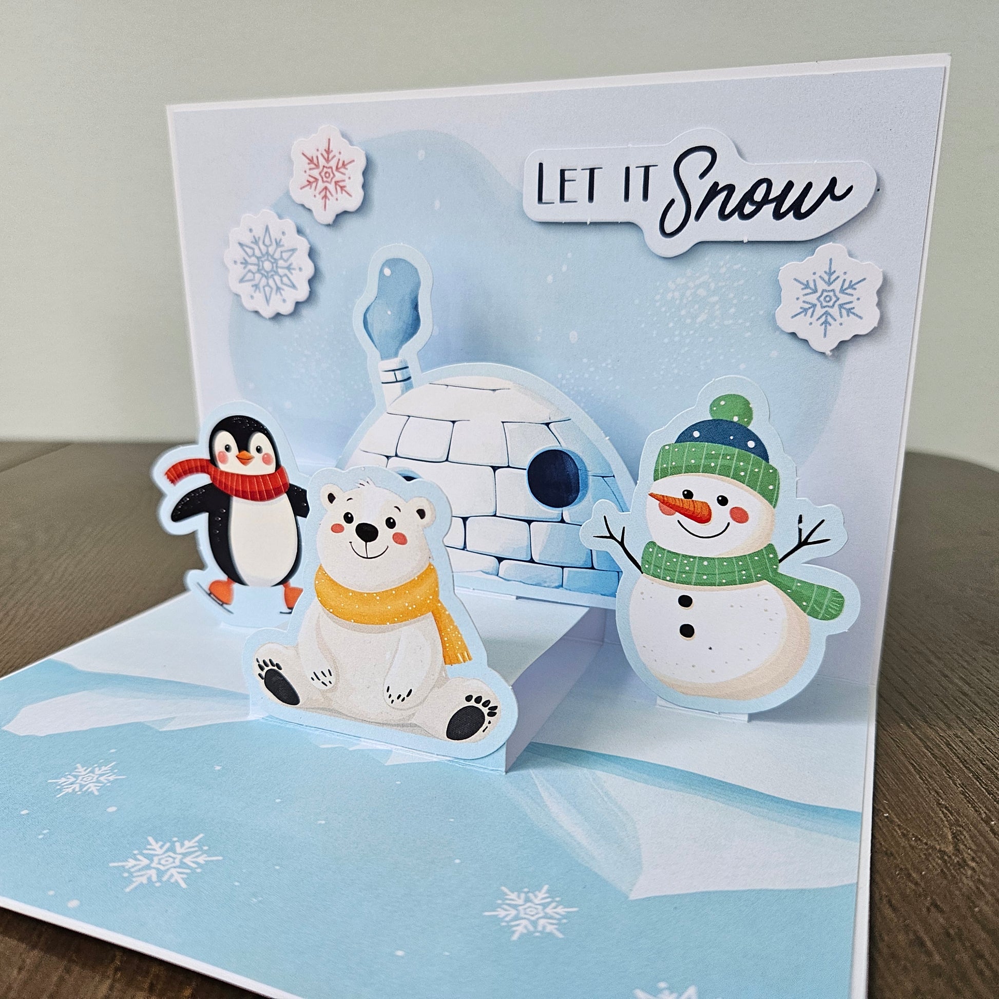 A handmade 3D pop-up holiday card featuring a snowy arctic scene with a polar bear, penguin, snowman, and igloo. The card has a white and light blue background with the text 'LET IT Snow' and decorative snowflakes.