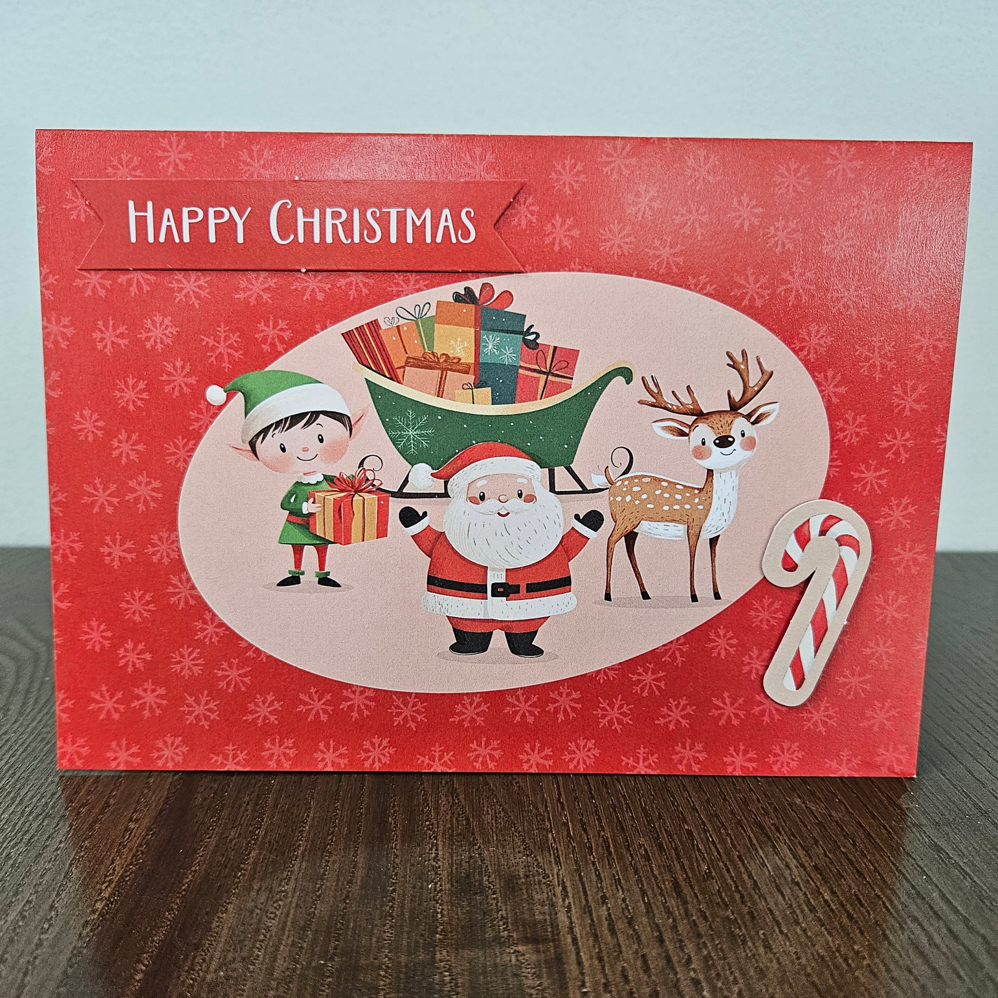 A red Christmas greeting card with 'Happy Christmas' at the top, featuring a festive illustration of Santa Claus, an elf, a reindeer, a candy cane, and a sleigh on a light wood surface.