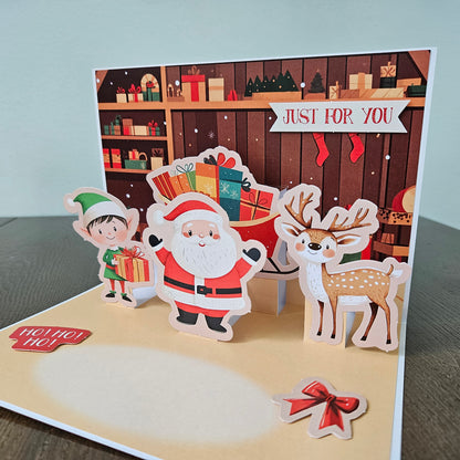 A 3D pop-up Christmas greeting card displayed on a dark wooden table against a light gray wall. The card features a festive holiday scene with a Santa Claus, an elf, a reindeer, and a gift box, with a wooden shelf background and a banner reading 'JUST FOR YOU'.