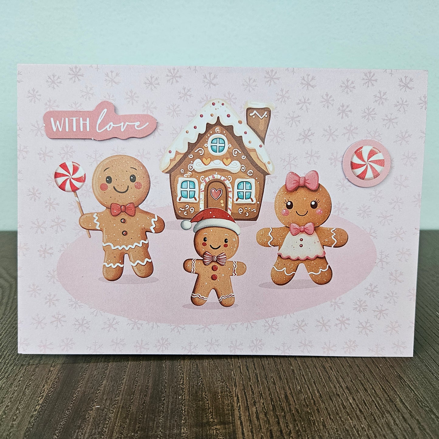 A square greeting card featuring a cute gingerbread holiday scene with three gingerbread figures, a gingerbread house, and a peppermint candy, displayed on a dark wooden surface against a light blue-green background.