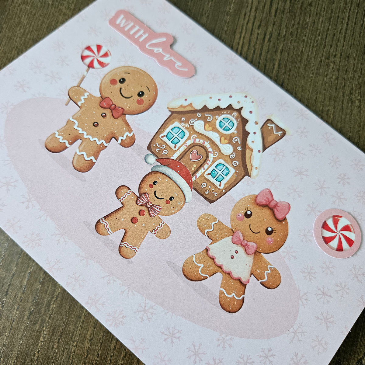A square decorative card or print featuring three gingerbread figures, a gingerbread house, a peppermint candy, and the phrase “with love,” photographed on a wood surface.