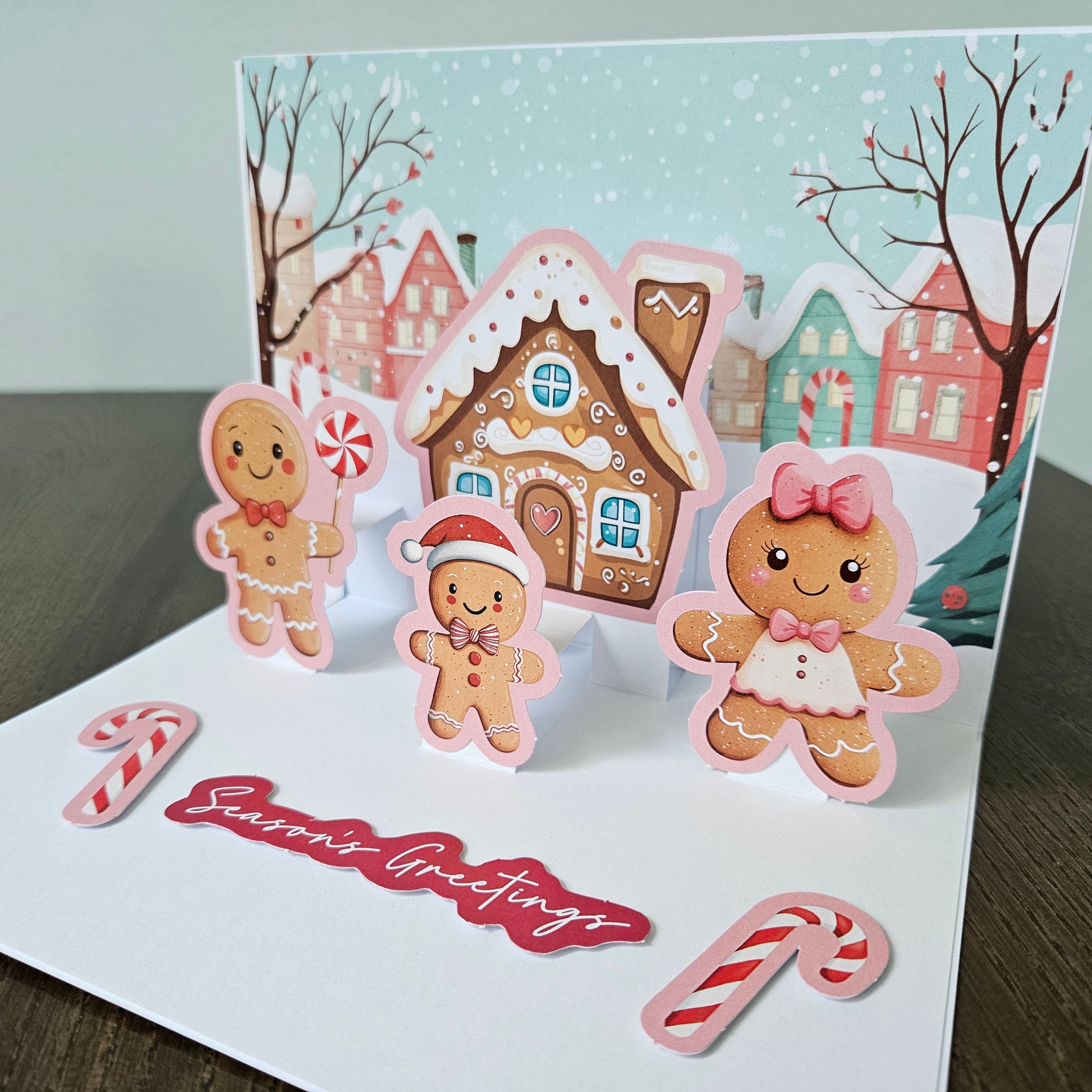 A handmade pop-up Christmas greeting card displayed on a dark wooden surface. The card features a snowy village scene with gingerbread houses and cookies, including three gingerbread men and a red script 'Season's Greetings' banner.