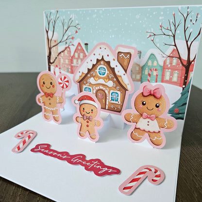 A handmade pop-up Christmas greeting card displayed on a dark wooden surface. The card features a snowy village scene with gingerbread houses and cookies, including three gingerbread men and a red script 'Season's Greetings' banner.