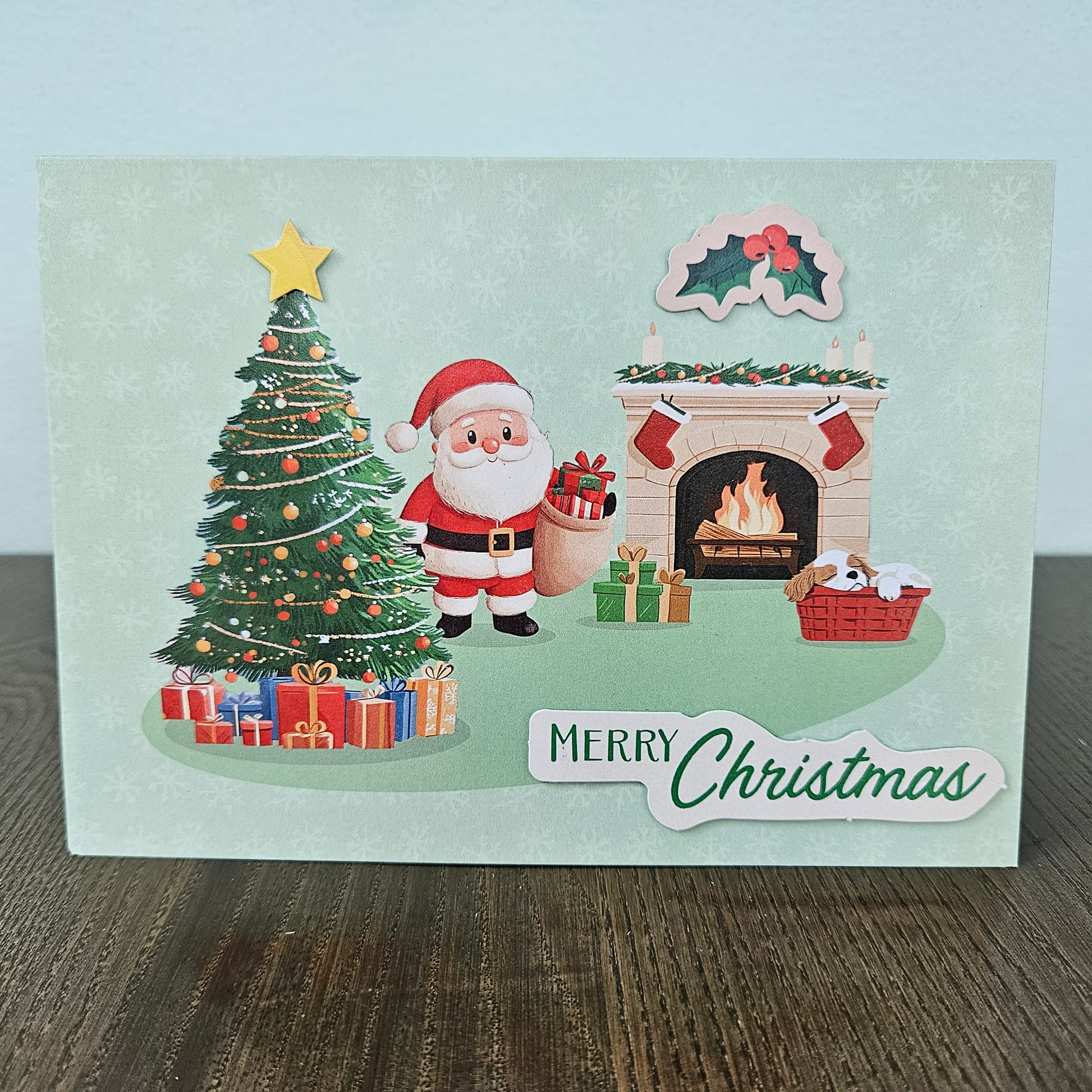 A Christmas greeting card featuring an illustrated Santa Claus standing beside a decorated Christmas tree, gifts, a burning fireplace, and a holly motif, with the text 'Merry Christmas' at the bottom. The card is photographed standing on a wooden surface against a pale wall.