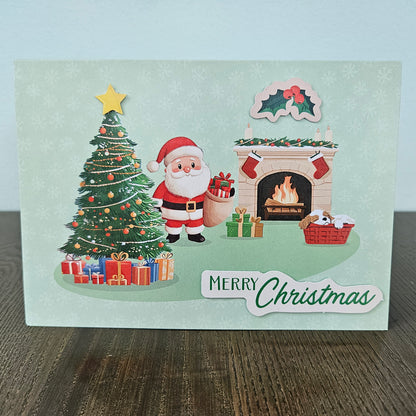 A Christmas greeting card featuring an illustrated Santa Claus standing beside a decorated Christmas tree, gifts, a burning fireplace, and a holly motif, with the text 'Merry Christmas' at the bottom. The card is photographed standing on a wooden surface against a pale wall.