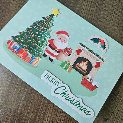 A handmade Christmas card with a light mint green background, featuring a colorful illustration of Santa Claus by a decorated Christmas tree and a burning fireplace, with a 'Merry Christmas' paper cutout on a wooden surface.