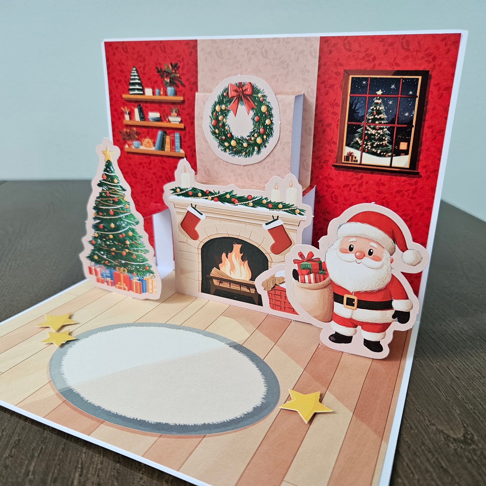 A 3D pop-up Christmas greeting card displayed on a dark wooden table. The card features a festive interior scene with a fireplace, a decorated Christmas tree, Santa Claus, a wreath, stars, and a blank oval space on the floor.