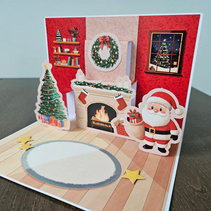A 3D pop-up Christmas greeting card displayed on a dark wooden table. The card features a festive interior scene with a fireplace, a decorated Christmas tree, Santa Claus, a wreath, stars, and a blank oval space on the floor.