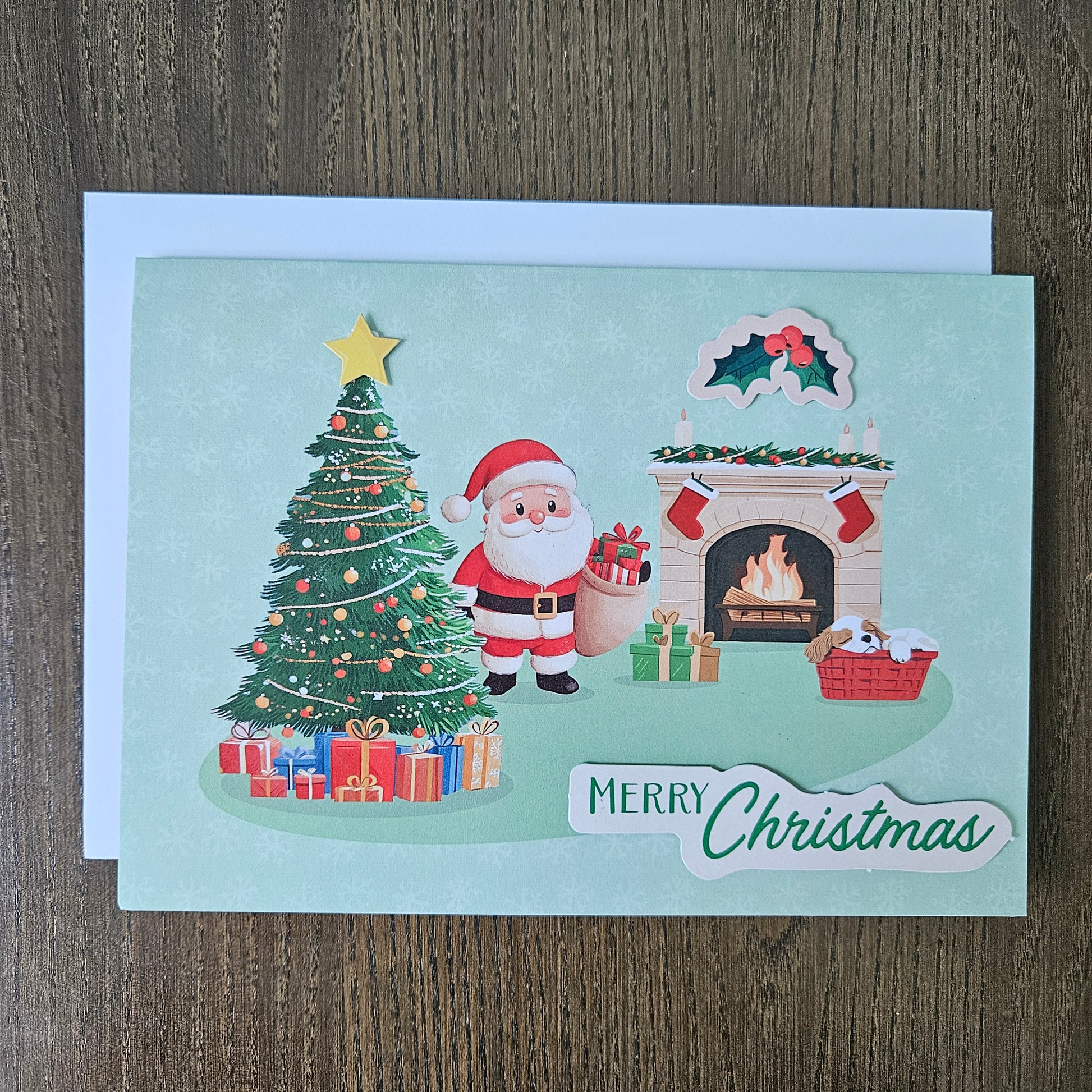 A Christmas greeting card featuring a festive illustration of Santa Claus by a decorated tree, gifts, a fireplace, and a 'Merry Christmas' banner, photographed on a wooden surface with a white envelope behind it.
