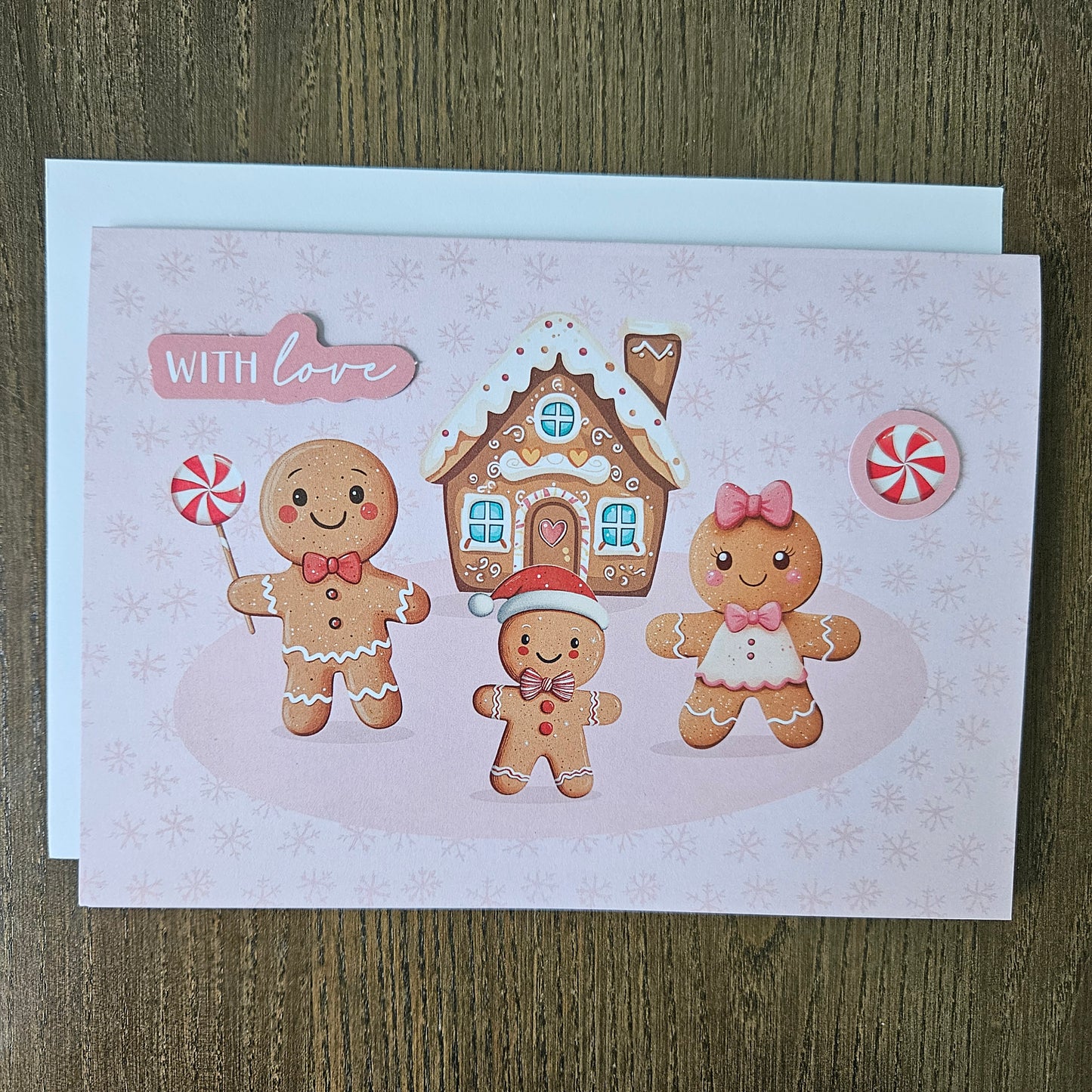 A square greeting card featuring cute gingerbread characters, a gingerbread house, candy canes, and the words 'WITH LOVE' on a pale pink background, placed on top of a white envelope over a wood surface.