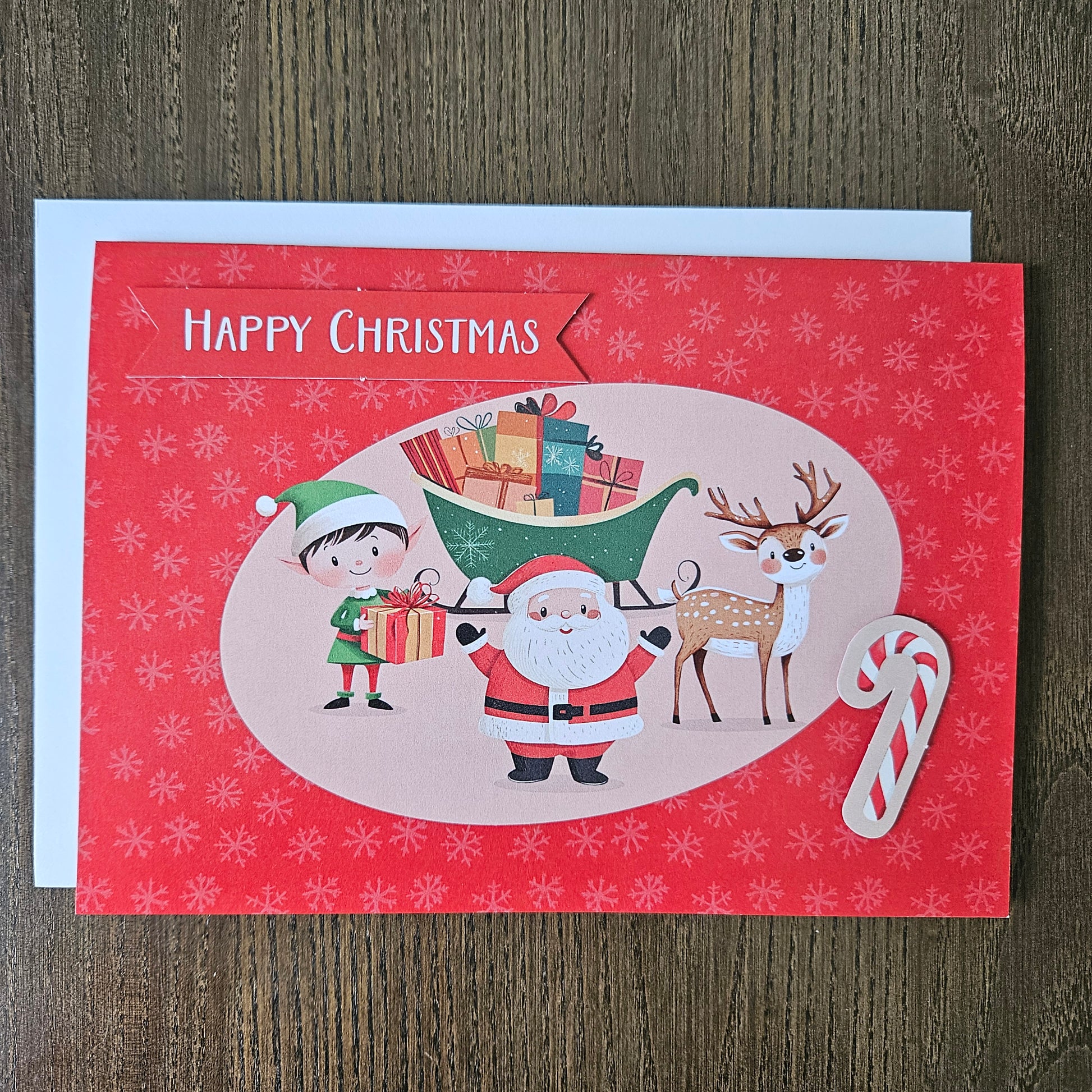 A red Christmas greeting card with a white envelope, placed on a brown wooden surface. The card features a festive illustration of Santa Claus, an elf, a reindeer, wrapped gifts, and a candy cane, with 'HAPPY CHRISTMAS' printed vertically on the left side.