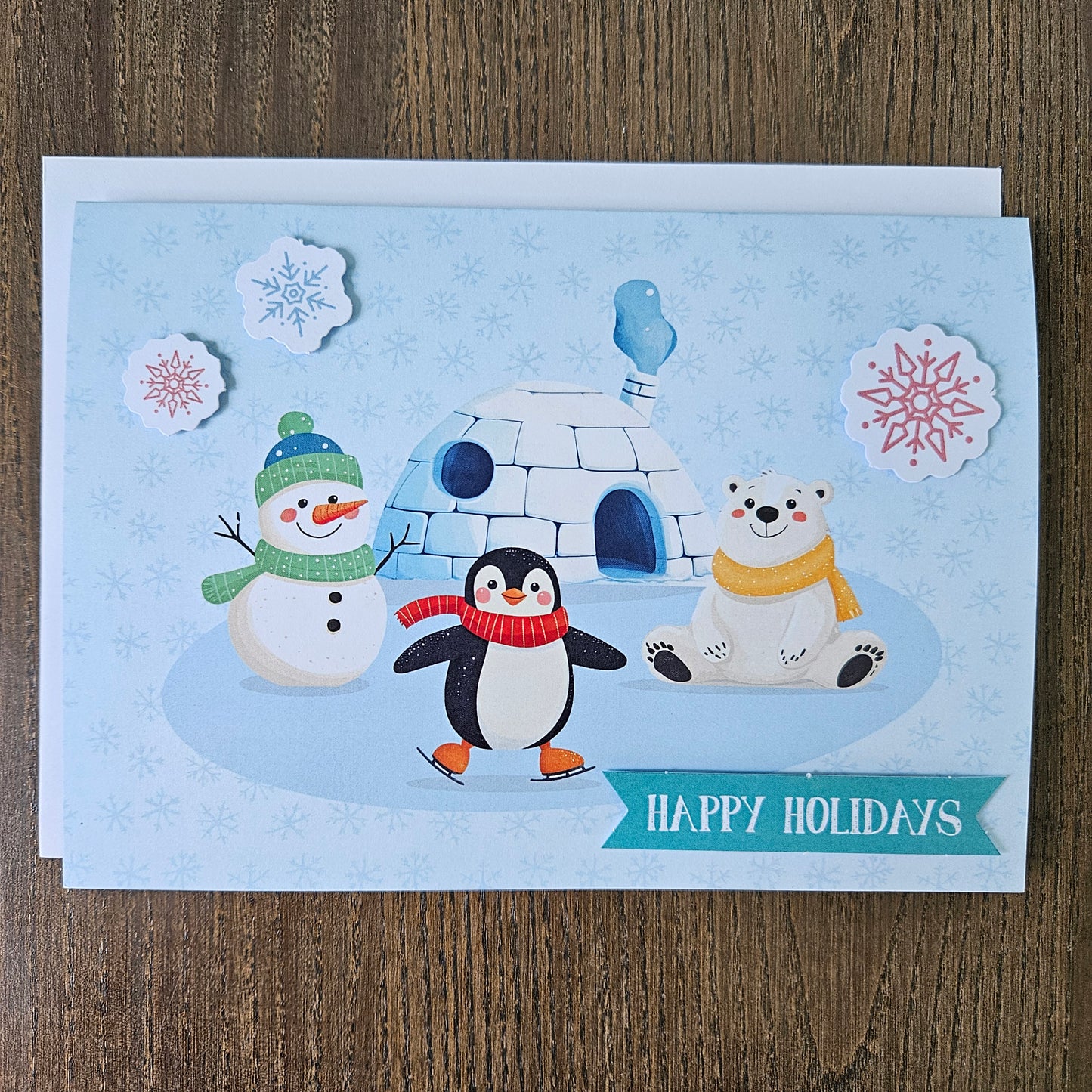 A square holiday greeting card featuring a winter scene with a snowman, penguin, polar bear, igloo, and snowflakes on a light blue background, placed on a wooden surface. A teal banner at the bottom reads “HAPPY HOLIDAYS.