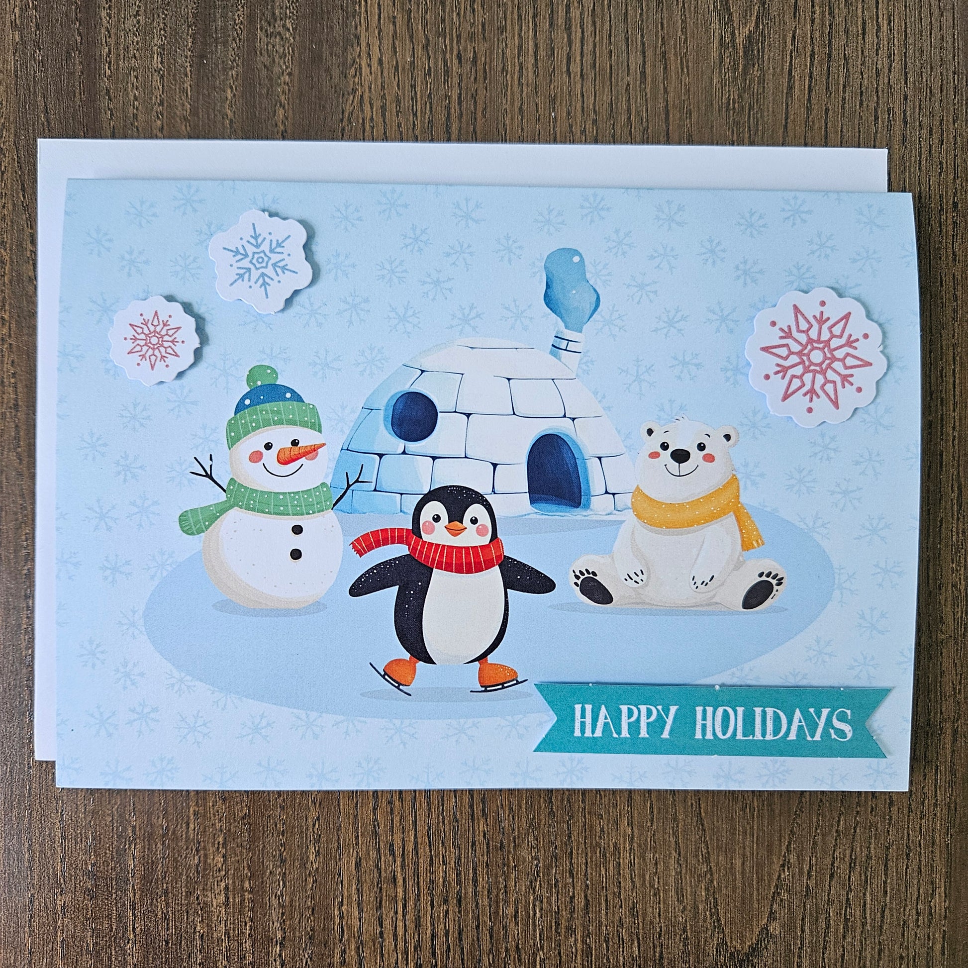 A square holiday greeting card featuring a winter scene with a snowman, penguin, polar bear, igloo, and snowflakes on a light blue background, placed on a wooden surface. A teal banner at the bottom reads “HAPPY HOLIDAYS.