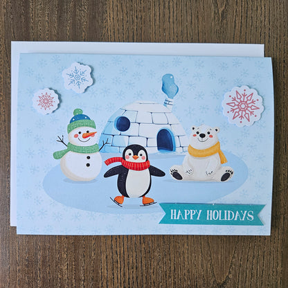 A square holiday greeting card featuring a winter scene with a snowman, penguin, polar bear, igloo, and snowflakes on a light blue background, placed on a wooden surface. A teal banner at the bottom reads “HAPPY HOLIDAYS.