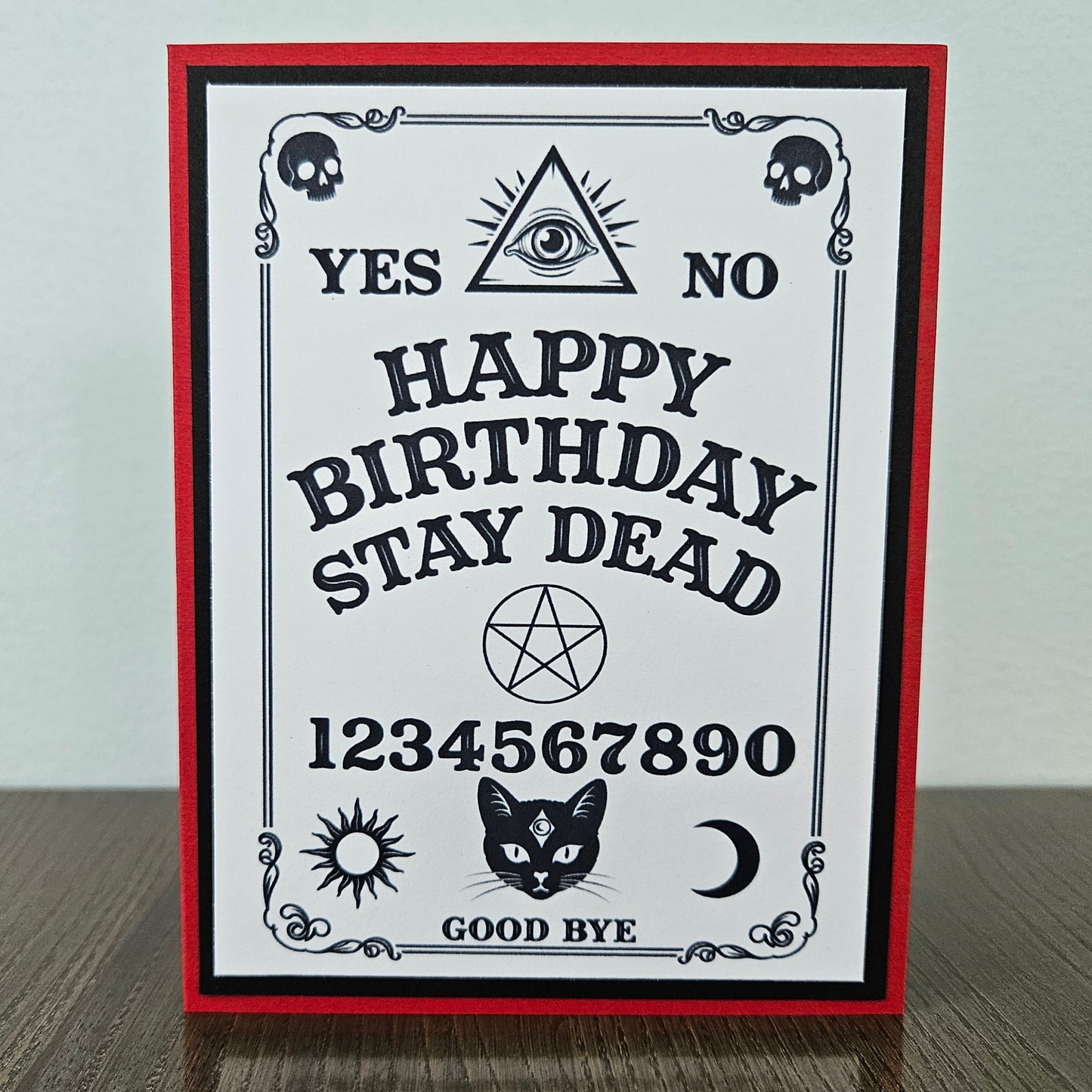 handmade A2 gothic birthday card featuring a black and white spirit board layout framed on a bold red cardstock base, with text reading Happy Birthday Stay Dead along with skulls, an eye of providence, a pentagram, and a black cat face with a third eye symbol.