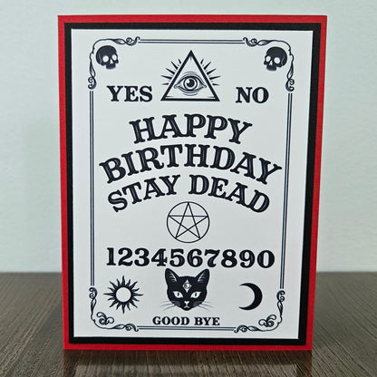 handmade A2 gothic birthday card featuring a black and white spirit board layout framed on a bold red cardstock base, with text reading Happy Birthday Stay Dead along with skulls, an eye of providence, a pentagram, and a black cat face with a third eye symbol.