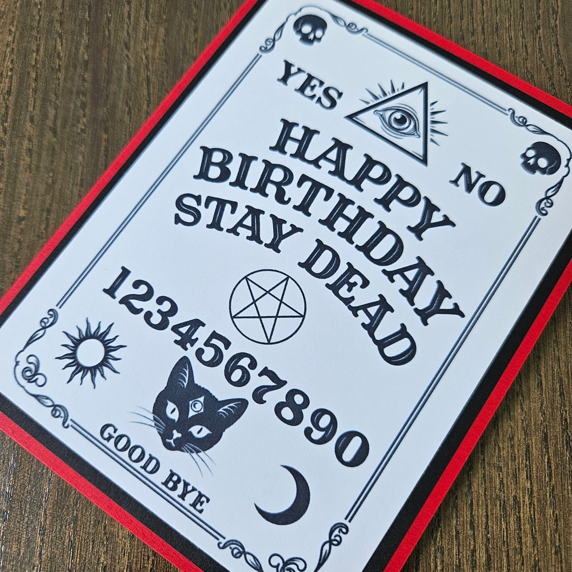 A gothic-style birthday card featuring a Ouija board design with the text 'YES NO HAPPY BIRTHDAY STAY DEAD 1234567890 GOOD BYE' along with illustrations of a cat, crescent moon, pentagram, eye, skulls, and sun, photographed on a wooden surface.