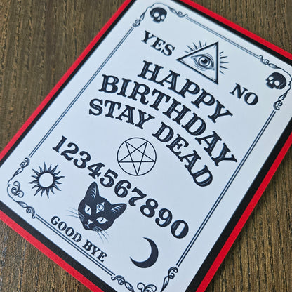 A gothic-style birthday card featuring a Ouija board design with the text 'YES NO HAPPY BIRTHDAY STAY DEAD 1234567890 GOOD BYE' along with illustrations of a cat, crescent moon, pentagram, eye, skulls, and sun, photographed on a wooden surface.