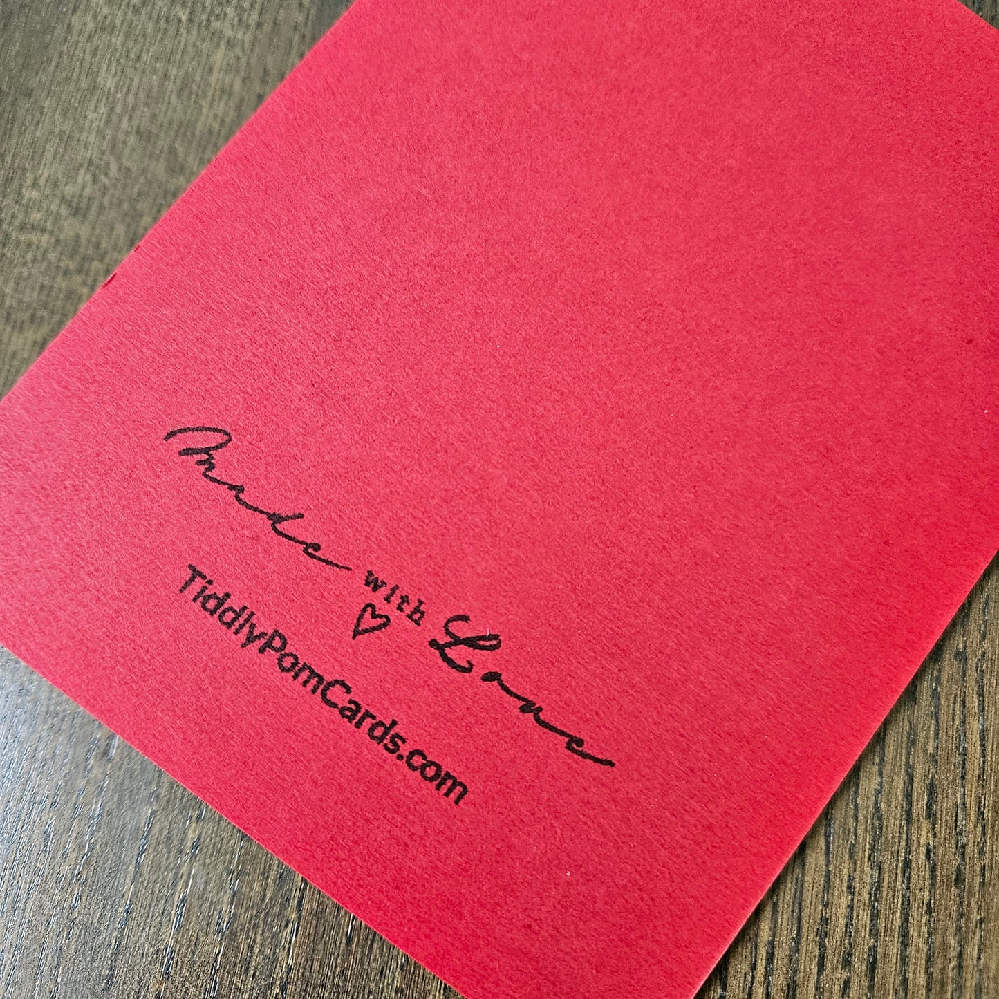 A close-up of a bright red textured card on a dark wood surface, printed with the words 'Proud with Love' and 'TiddlyPomCards.com' in black script.