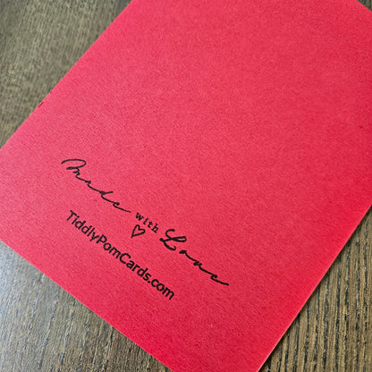 A close-up of a bright red textured card on a dark wood surface, printed with the words 'Proud with Love' and 'TiddlyPomCards.com' in black script.