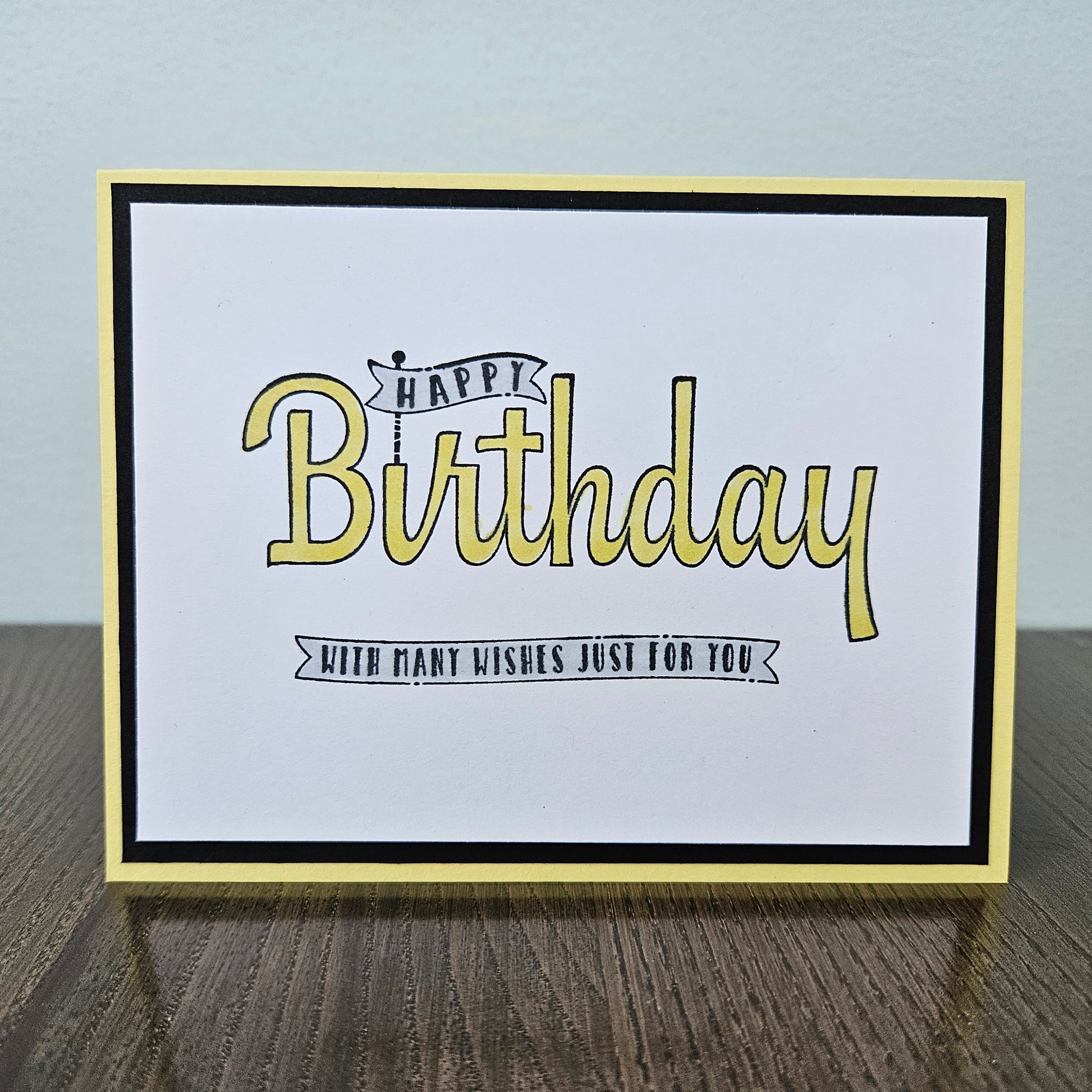 A square birthday greeting card with a white center, black and yellow border, and the words “Happy Birthday” in yellow script with black outlines, plus smaller text reading “With many wishes just for you,” shown standing on a wooden surface against a pale wall.