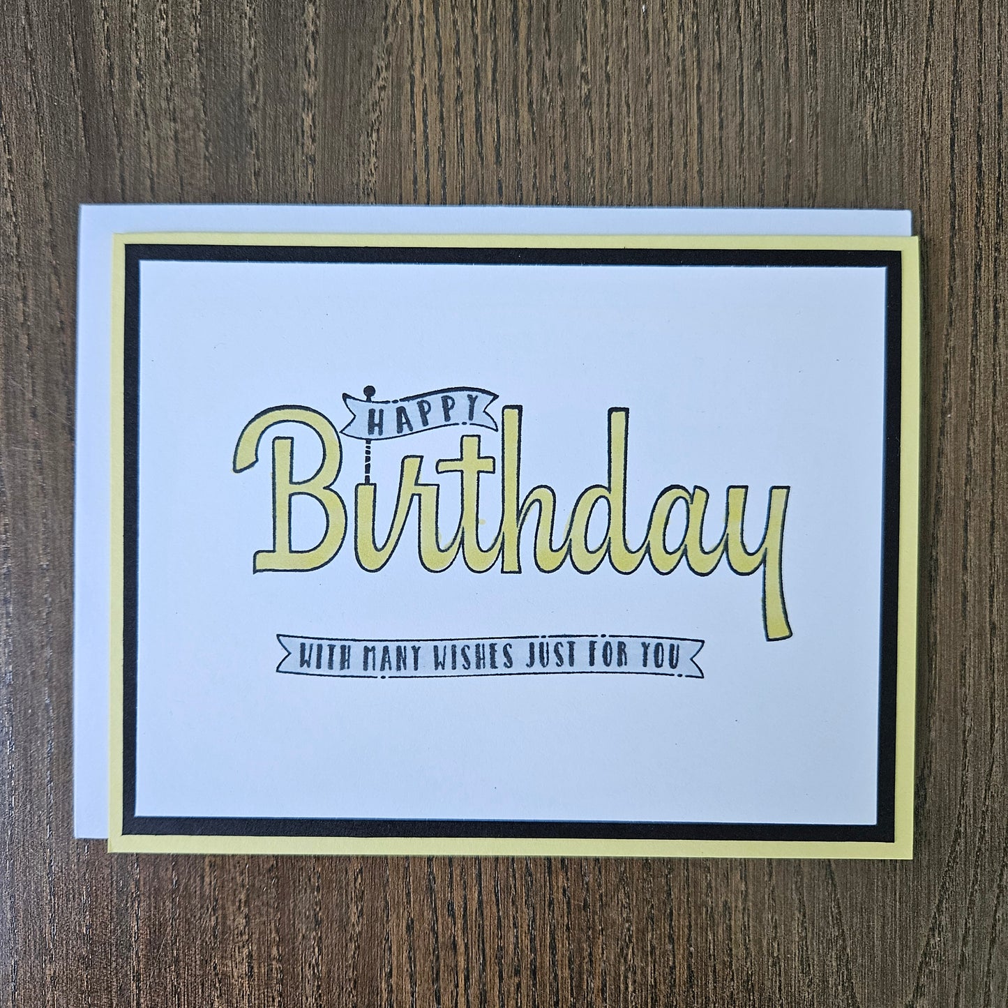 A square birthday card with a white center, black border, and pale yellow outer border, placed on a brown wood surface. The card reads “Happy Birthday” in large pale yellow script with smaller black text that says “With many wishes just for you.