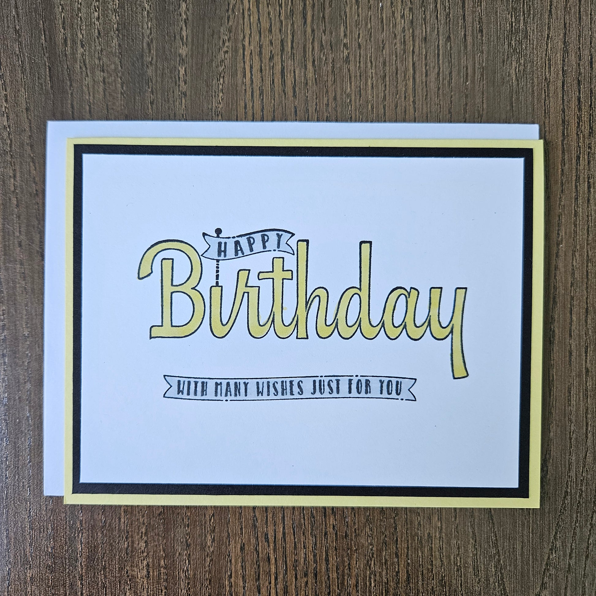 A square birthday card with a white center, black border, and pale yellow outer border, placed on a brown wood surface. The card reads “Happy Birthday” in large pale yellow script with smaller black text that says “With many wishes just for you.