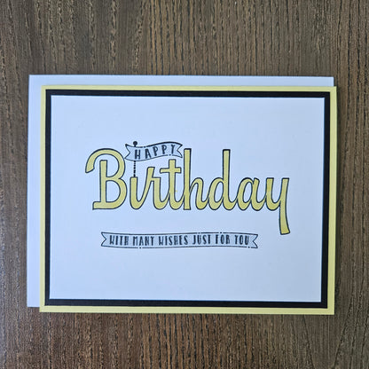 A square birthday card with a white center, black border, and pale yellow outer border, placed on a brown wood surface. The card reads “Happy Birthday” in large pale yellow script with smaller black text that says “With many wishes just for you.