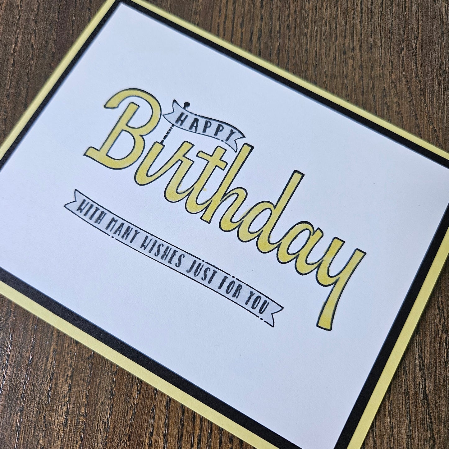 A square birthday card with a white center, black border, and pale yellow frame, placed on a brown wood surface. The card reads “Happy Birthday” in large yellow script with smaller black text that says “With kind wishes just for you.