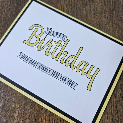 A square birthday card with a white center, black border, and pale yellow frame, placed on a brown wood surface. The card reads “Happy Birthday” in large yellow script with smaller black text that says “With kind wishes just for you.
